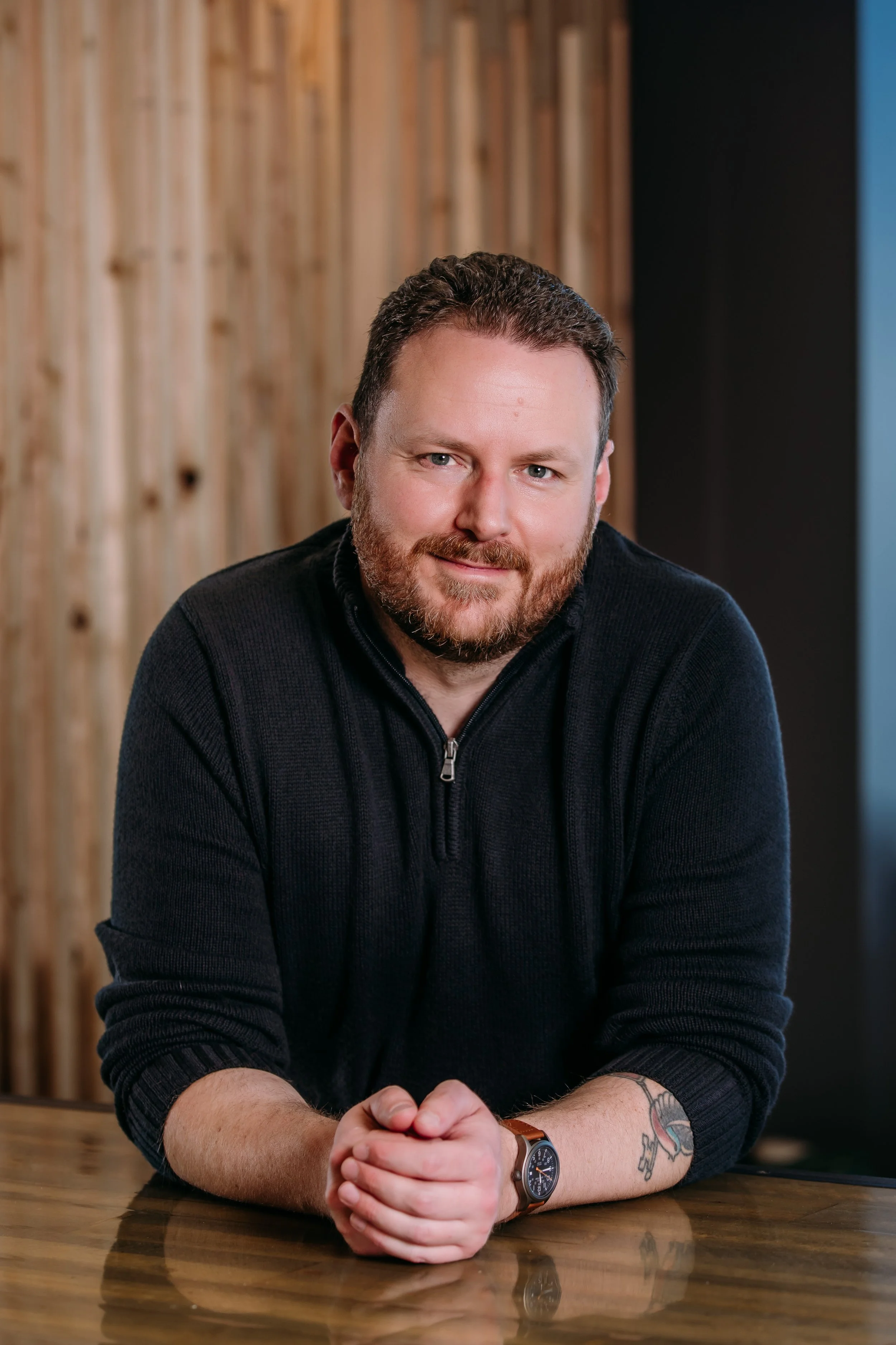 Jonathan P. Morrow wearing a black zip-up sweater, sitting at a wooden table in front of a wooden wall, looking at the camera.