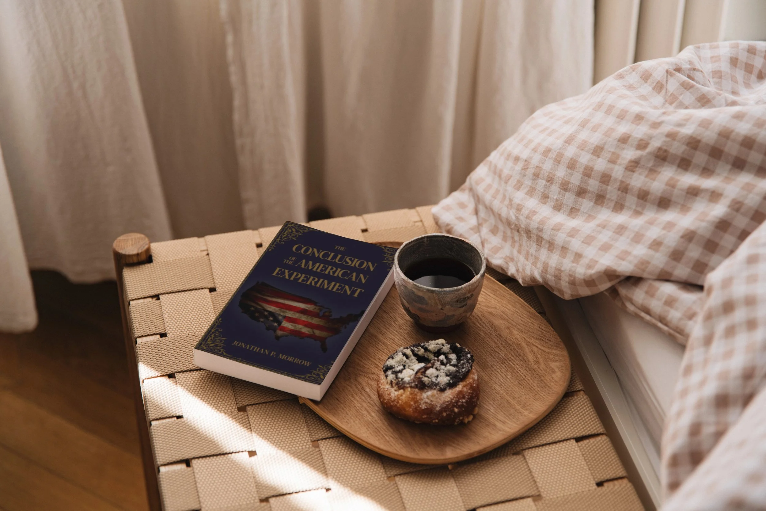 A bedside table with a book titled 'The Conclusion of the American Experiment,' a cup of coffee, and a pastry, next to a bed with checkered sheets.