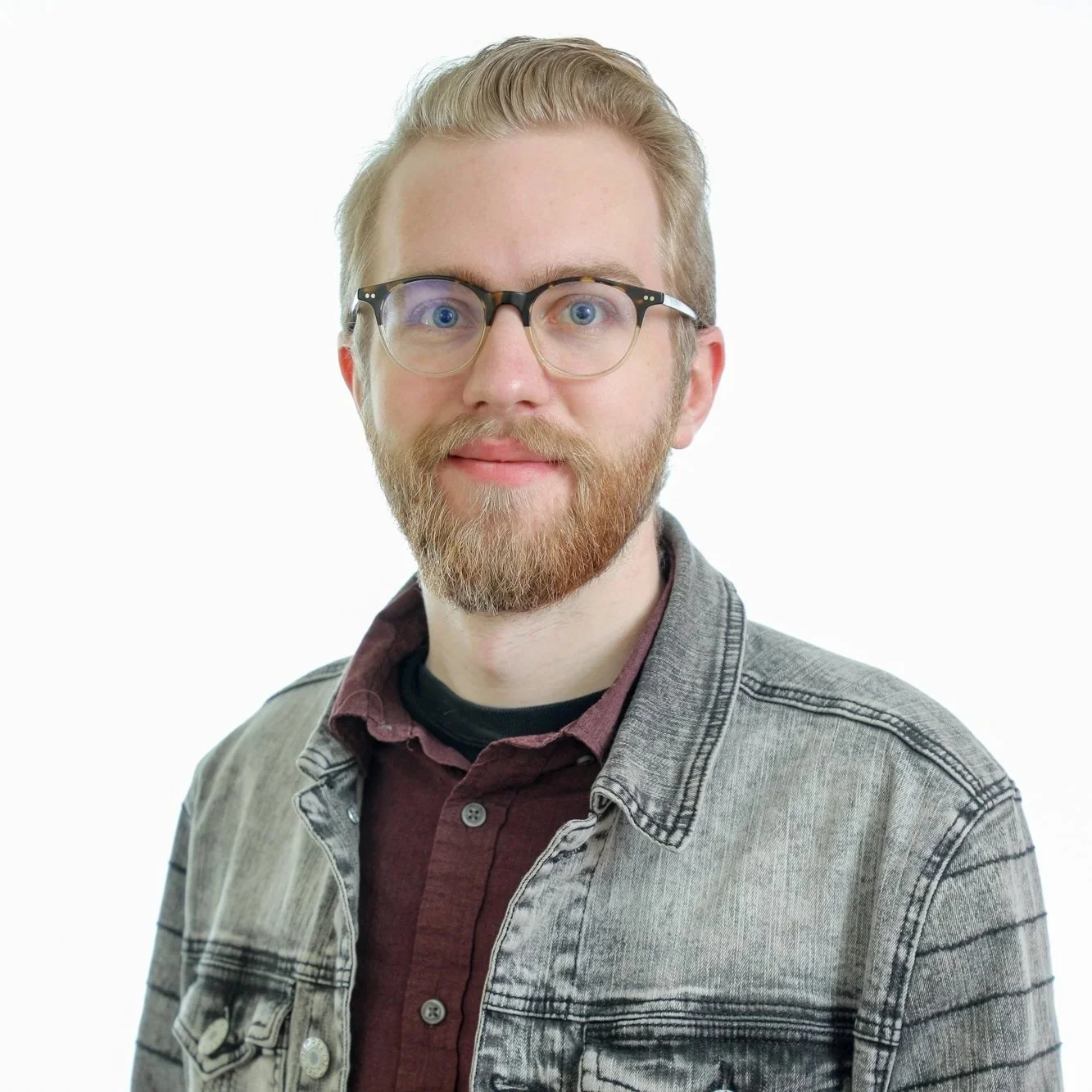 A young man with glasses, a beard, and mustache, wearing a denim jacket over a maroon shirt, standing against a white background.
