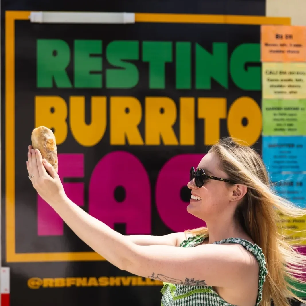 A woman smiling and taking a selfie with a burrito in front of a colorful sign that reads "Resting Burrito Face."