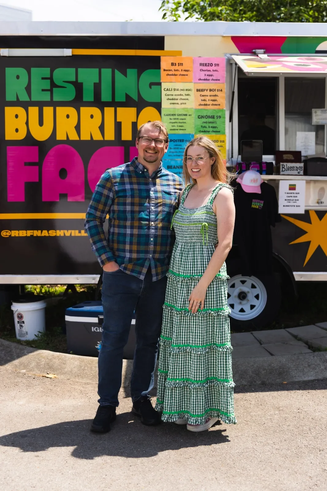 A couple standing in front of a food truck labeled 'Resting Burrito Face.' The man is wearing glasses, a plaid shirt, and jeans, and the woman is wearing glasses and a green and white checkered dress. The food truck menu is visible in the background.
