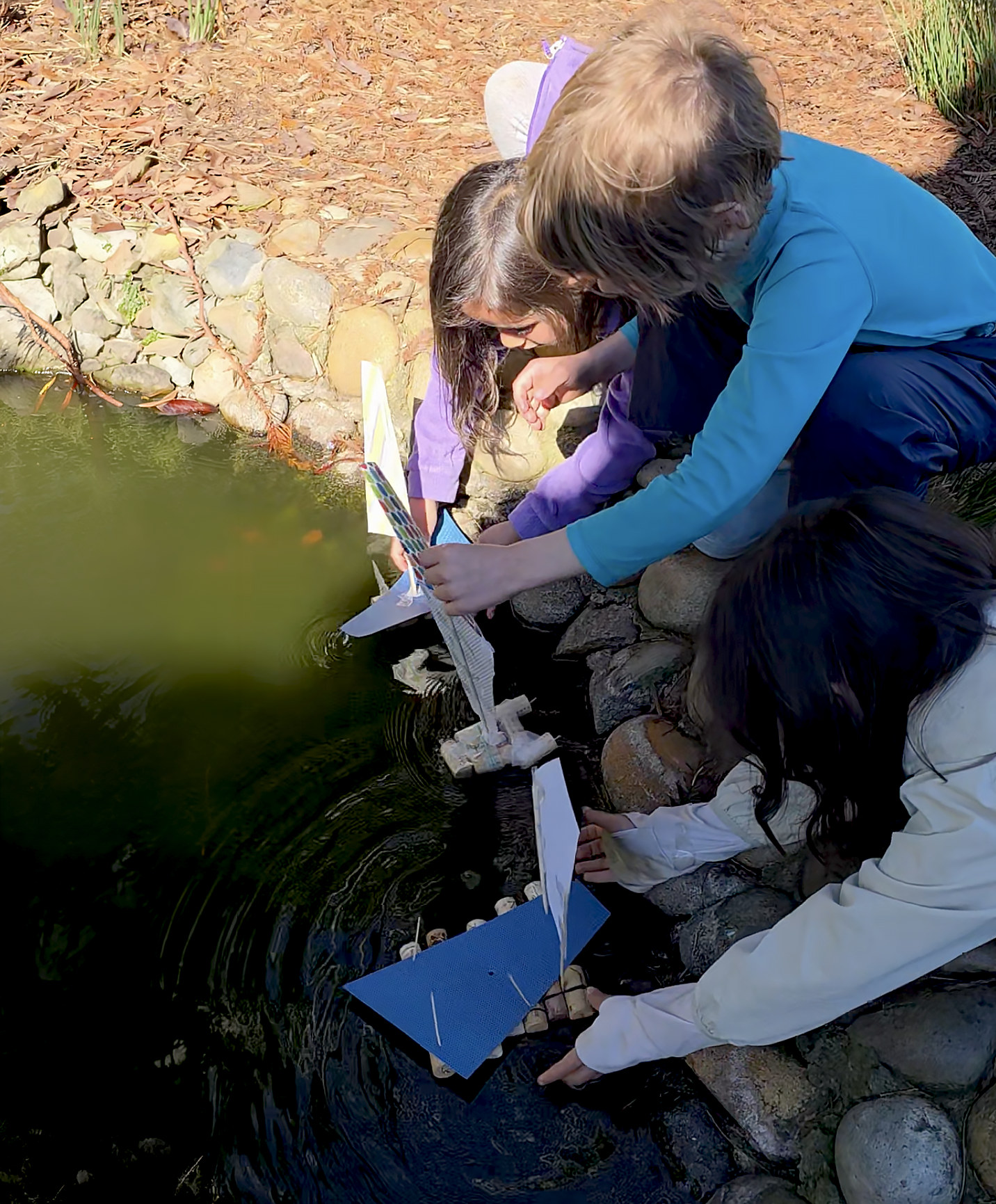 Three Sylva School students testing their handmade sailboats developed for their child-led interest projects, in a lake.