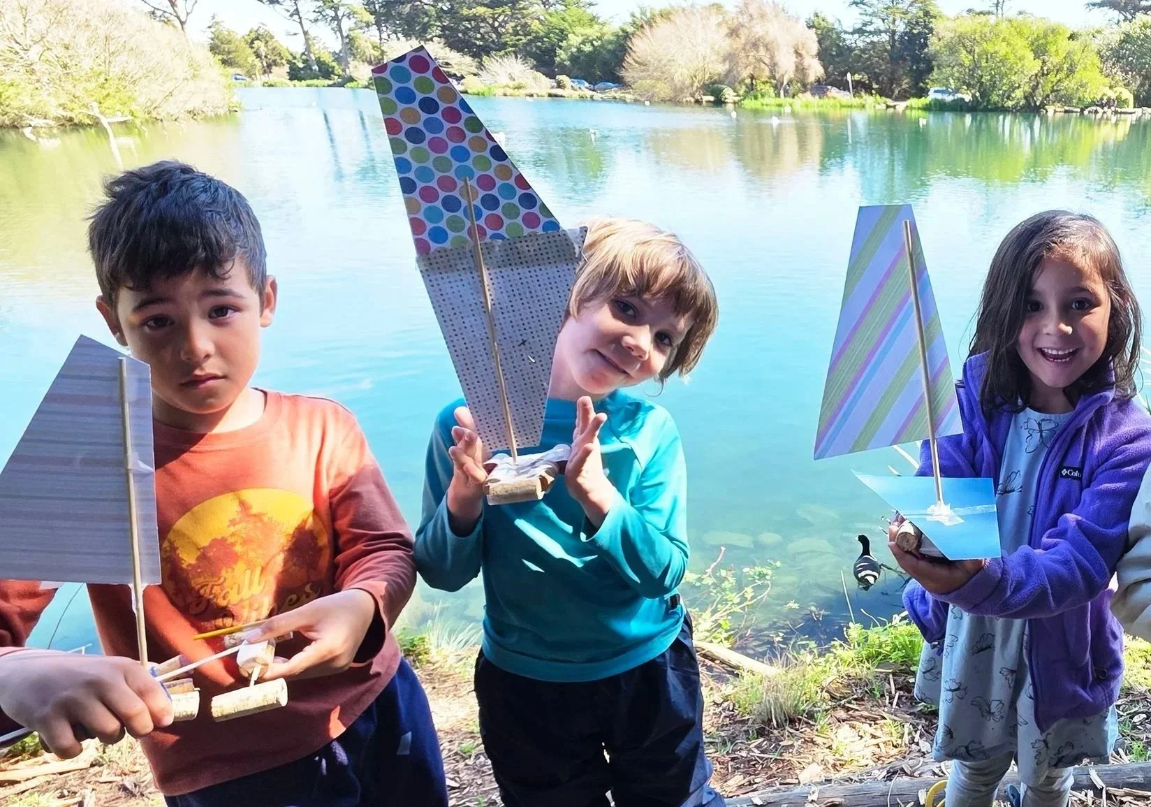Three Sylva School students holding handmade sailboats developed for their child-led interest projects, by a lake with trees and blue sky in the background.