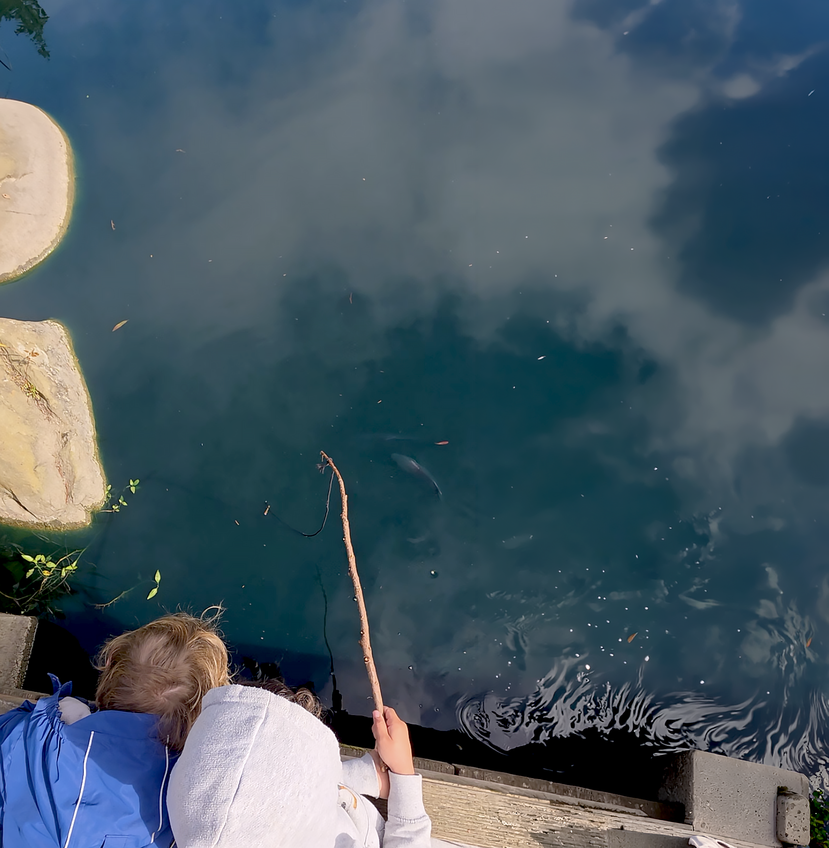 Two Sylva School students with a homemade finish pole, fishing off a stone dock into a lake surrounded by water with reflections of the sky and clouds.
