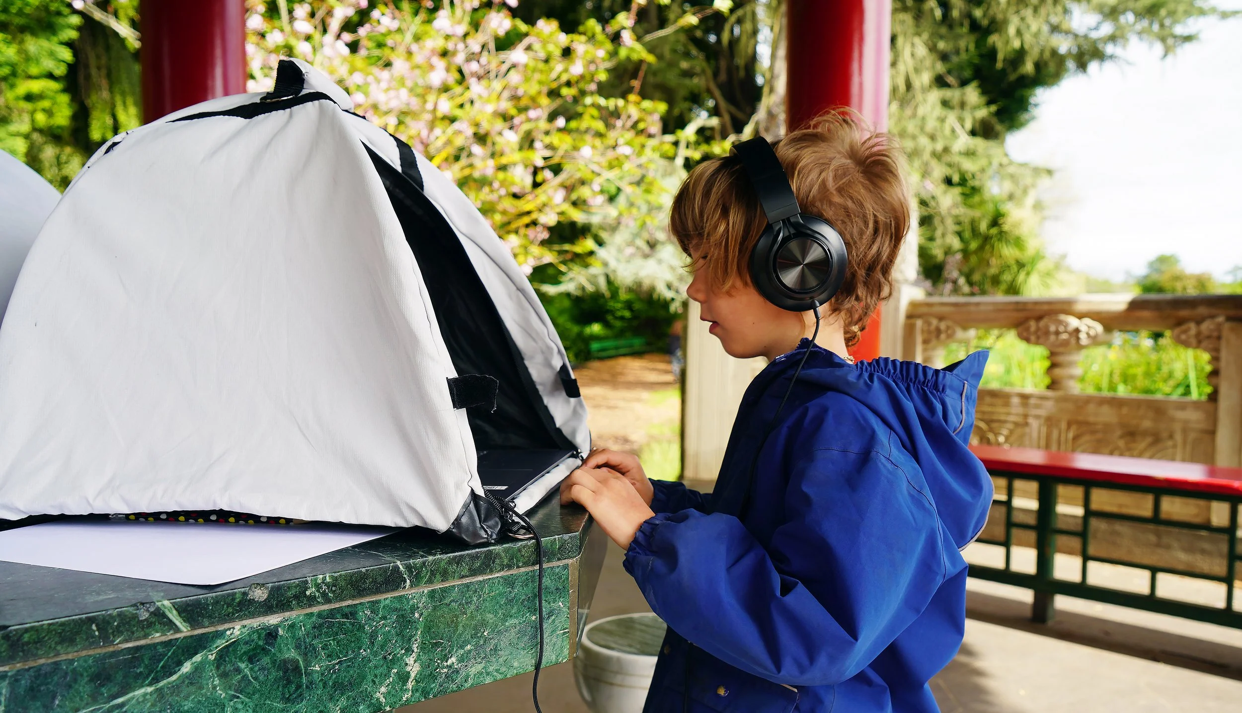 A Sylva School student with curly hair wearing a blue jacket and large black headphones, sitting at a green marble table under a pavilion outside, doing academics on a laptop set up inside a white tent, with trees and pink blossoms in the background.