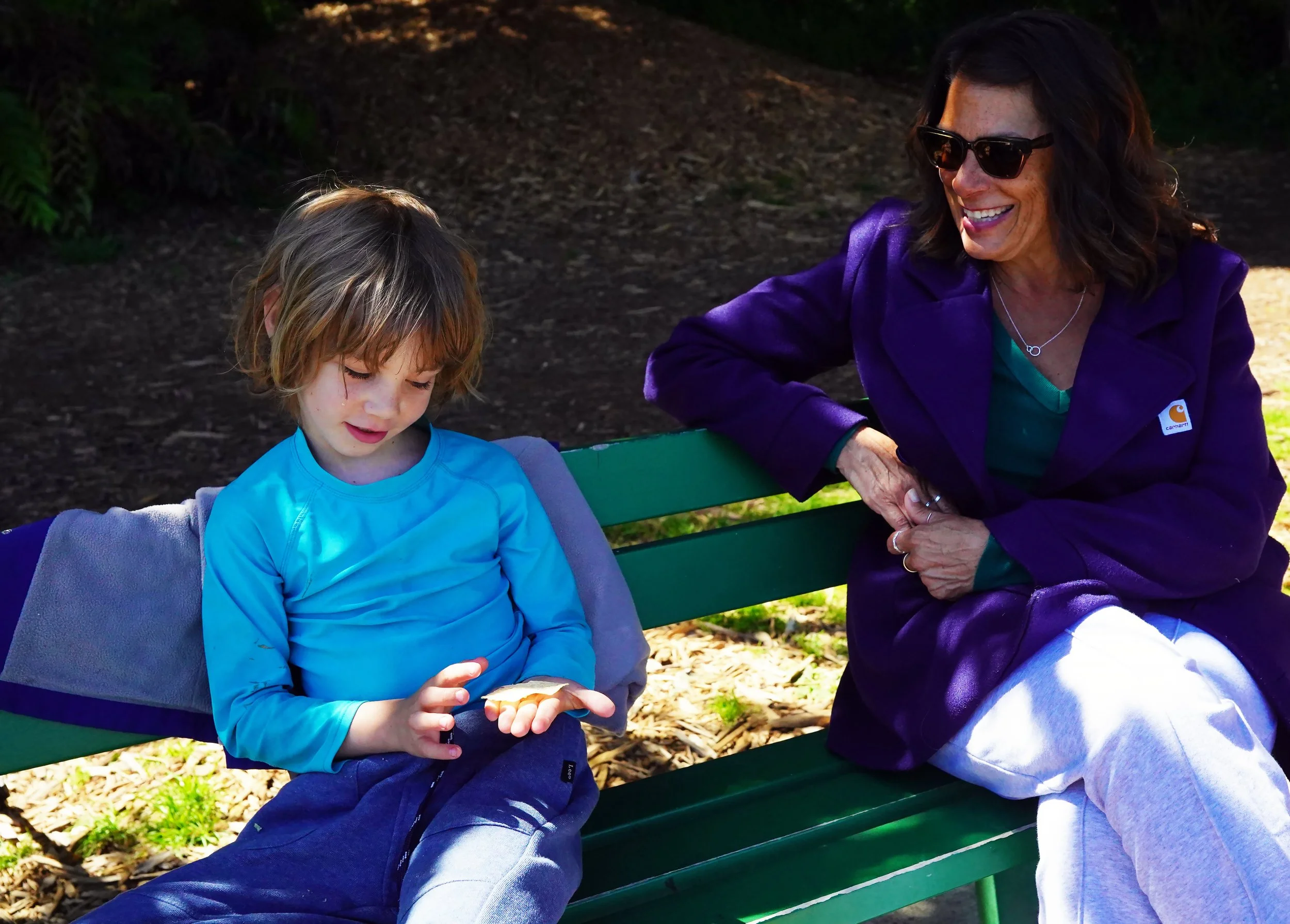 Sylva School Founder, Laura Engel, sitting on a park bench smiling at a student with light brown hair, who is looking down at a leaf in his hands, in a park setting with sunlight and trees in the background.