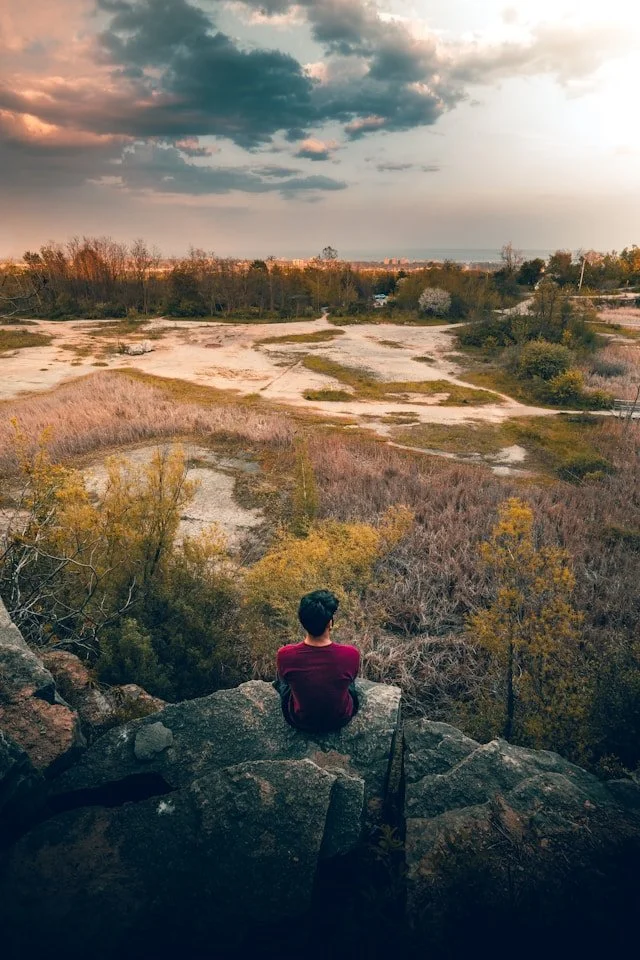 A person sitting on a rocky ledge overlooking a landscape with sparse trees, open land, and a cloudy sky at sunset.