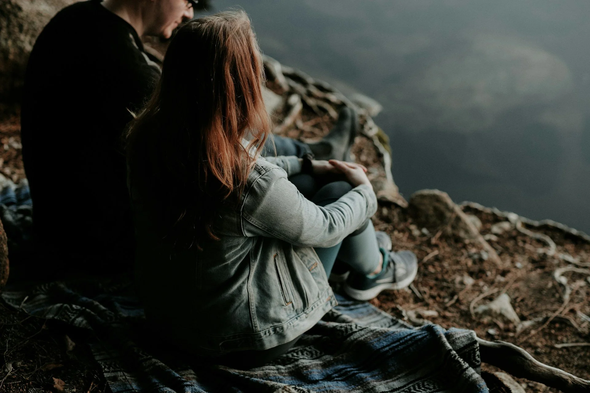 Two people sitting on a rocky shore by the water, looking out over a lake or river, with their hands clasped together.