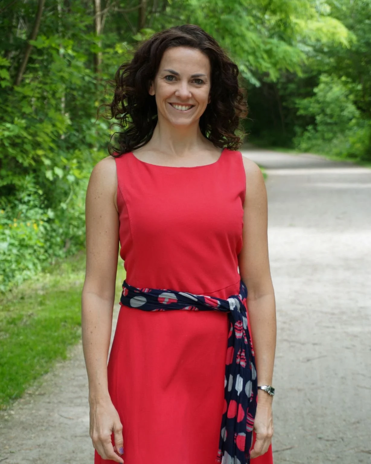 A woman with dark curly hair smiling outdoors on a forest path, wearing a sleeveless red dress with a blue patterned scarf tied around her waist.