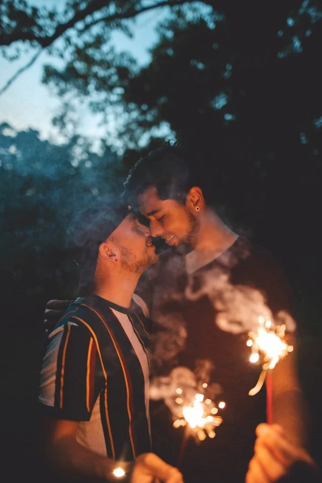 Two men standing close together at night, holding sparklers, with smoke and trees in the background.