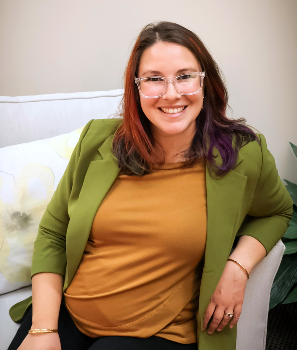 A woman with glasses, multicolored hair, and wearing a green blazer and gold top, sitting on a beige chair and smiling.