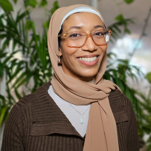 A smiling woman wearing glasses, a beige hijab, and a brown sweater stands in front of green plants.
