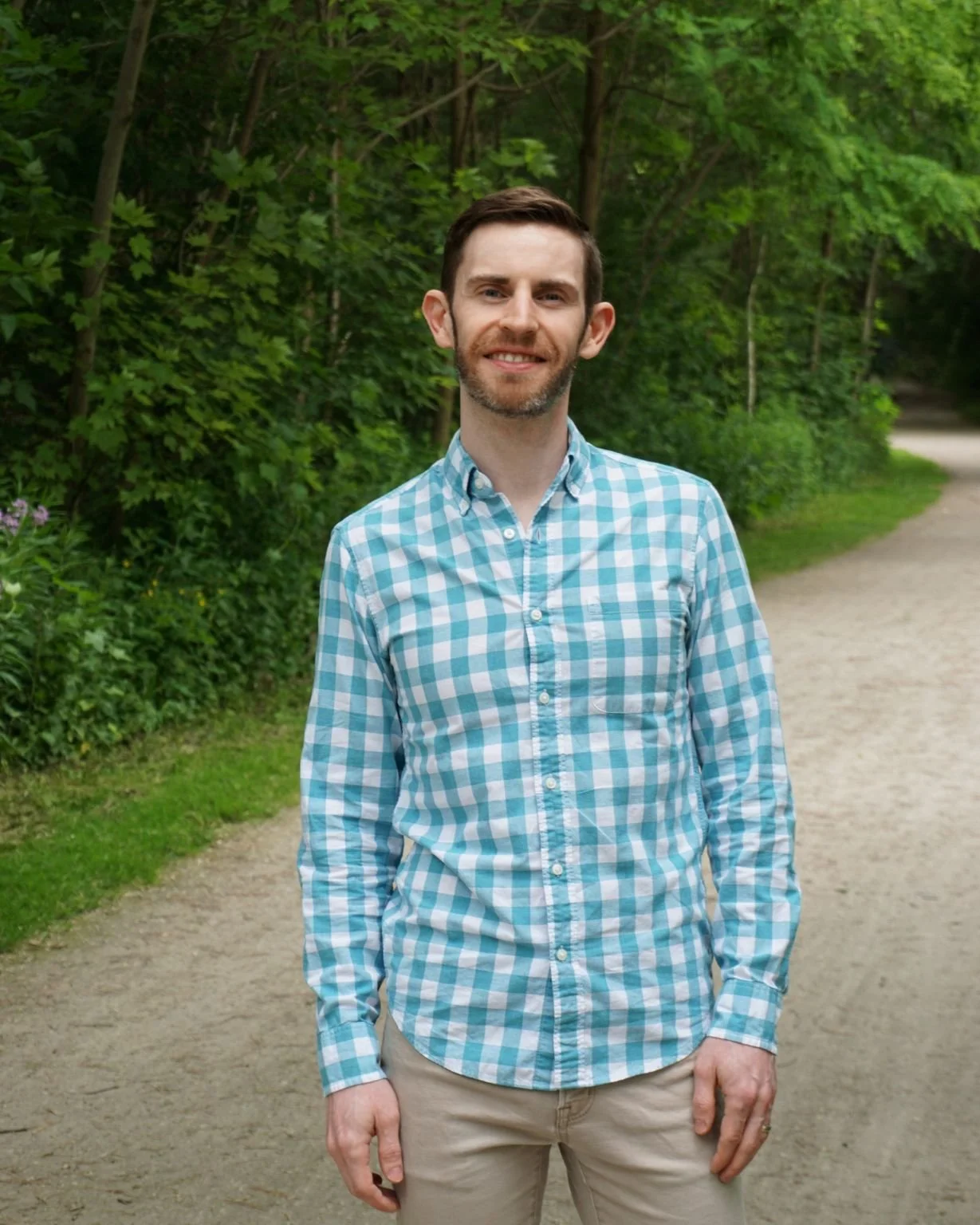 A man standing outdoors on a dirt path, wearing a light blue and white checkered shirt and beige pants, smiling with greenery in the background.