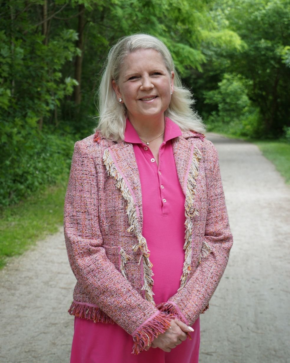 A woman with light blonde hair stands outdoors on a dirt path surrounded by green trees. She is wearing a pink top and a pink tweed jacket with fringes, smiling at the camera.