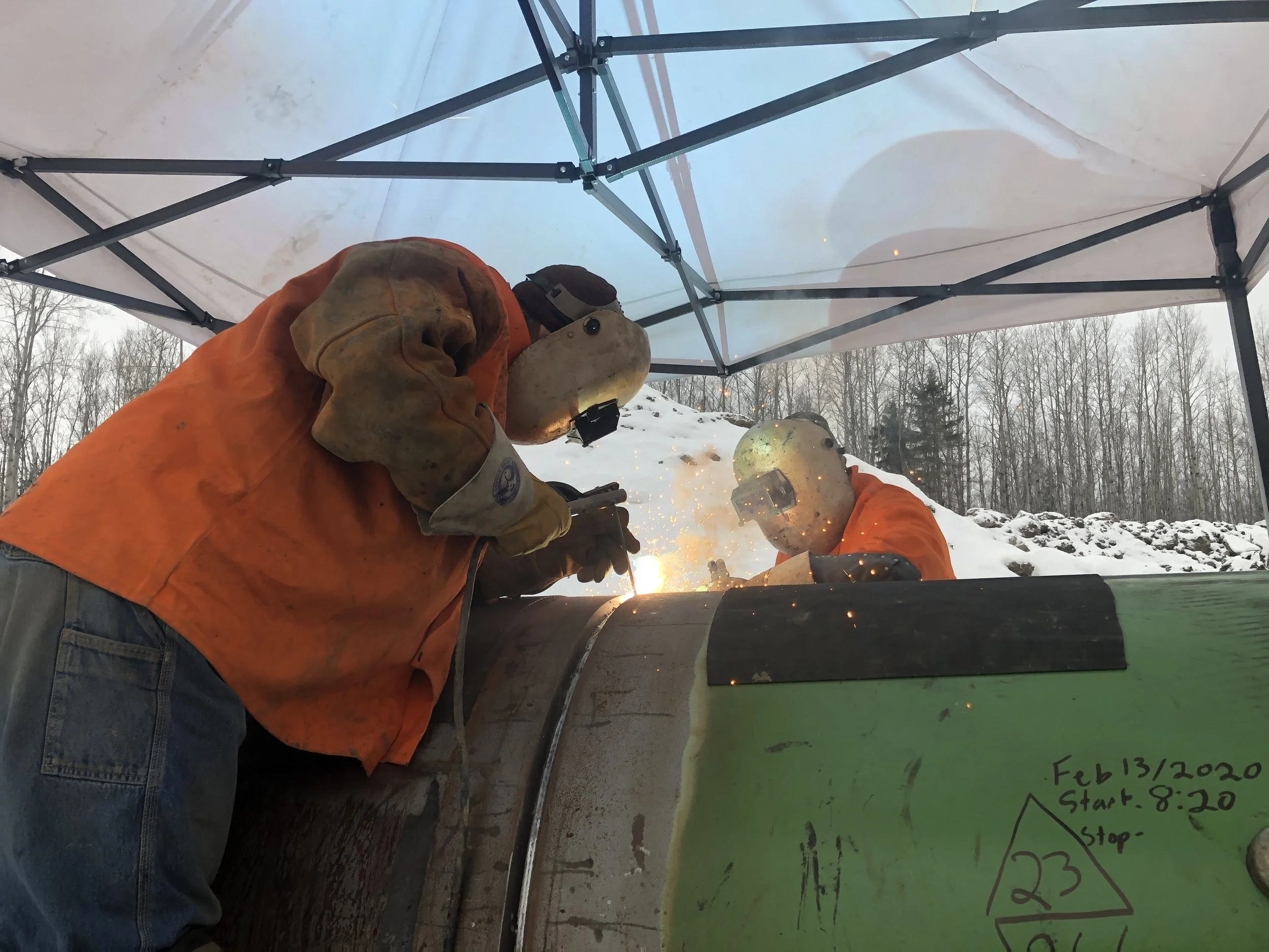 Two individuals wearing welding masks and orange protective jackets welding metal outdoors under a canopy with snow on the ground and leafless trees in the background.