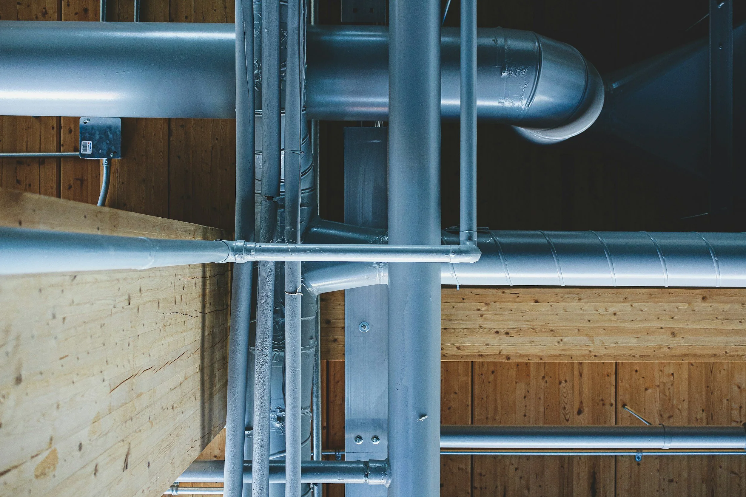 View of industrial metal pipes and ducts mounted on a wooden ceiling in an unfinished building.
