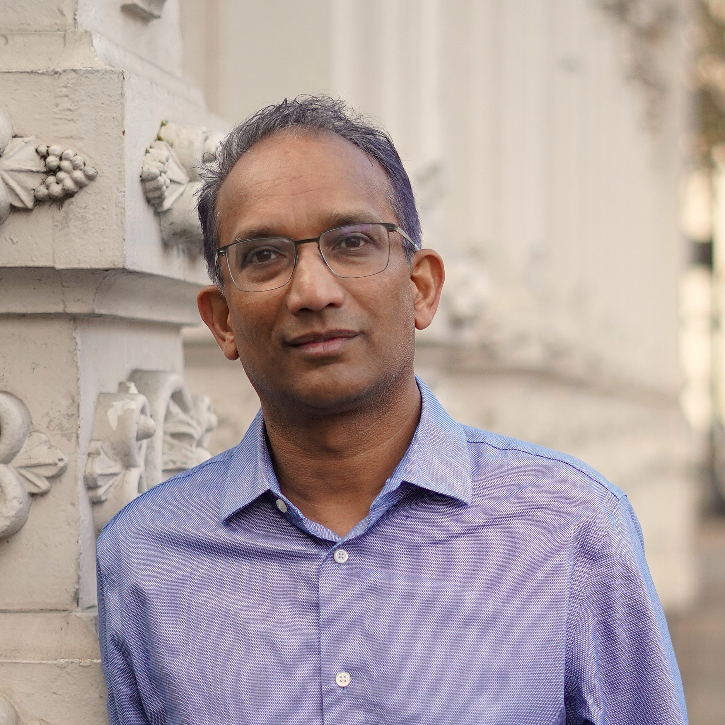 Partial view of a man wearing glasses and a blue button-up shirt, standing outdoors near a white decorative wall.