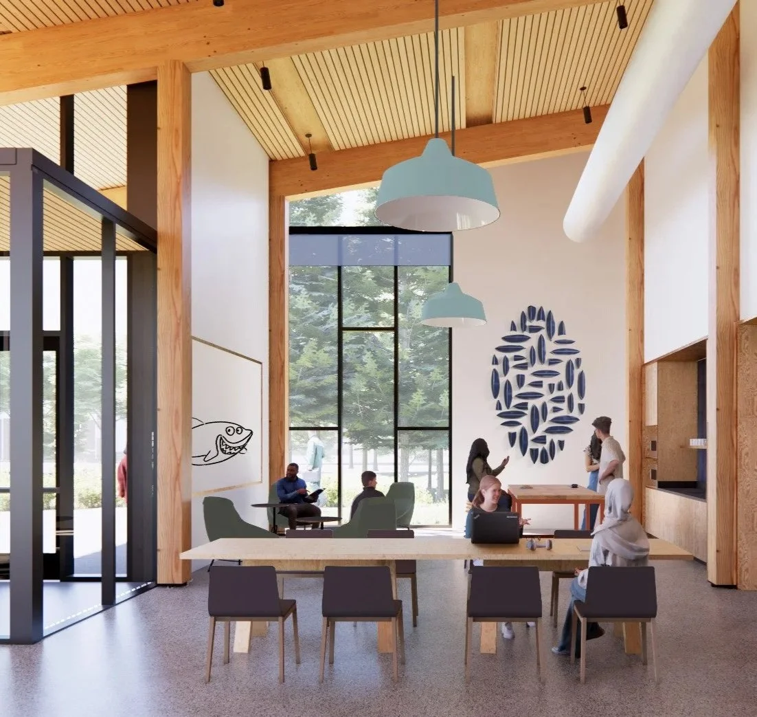 Modern indoor space with high ceilings featuring wooden beams and large windows showing trees outside. Multiple people are sitting and standing, engaging with electronic devices. Decorative art, including a stylized fish and an oval-shaped pattern, adorns the white walls. There's a large wooden table with chairs and hanging pendant lights.