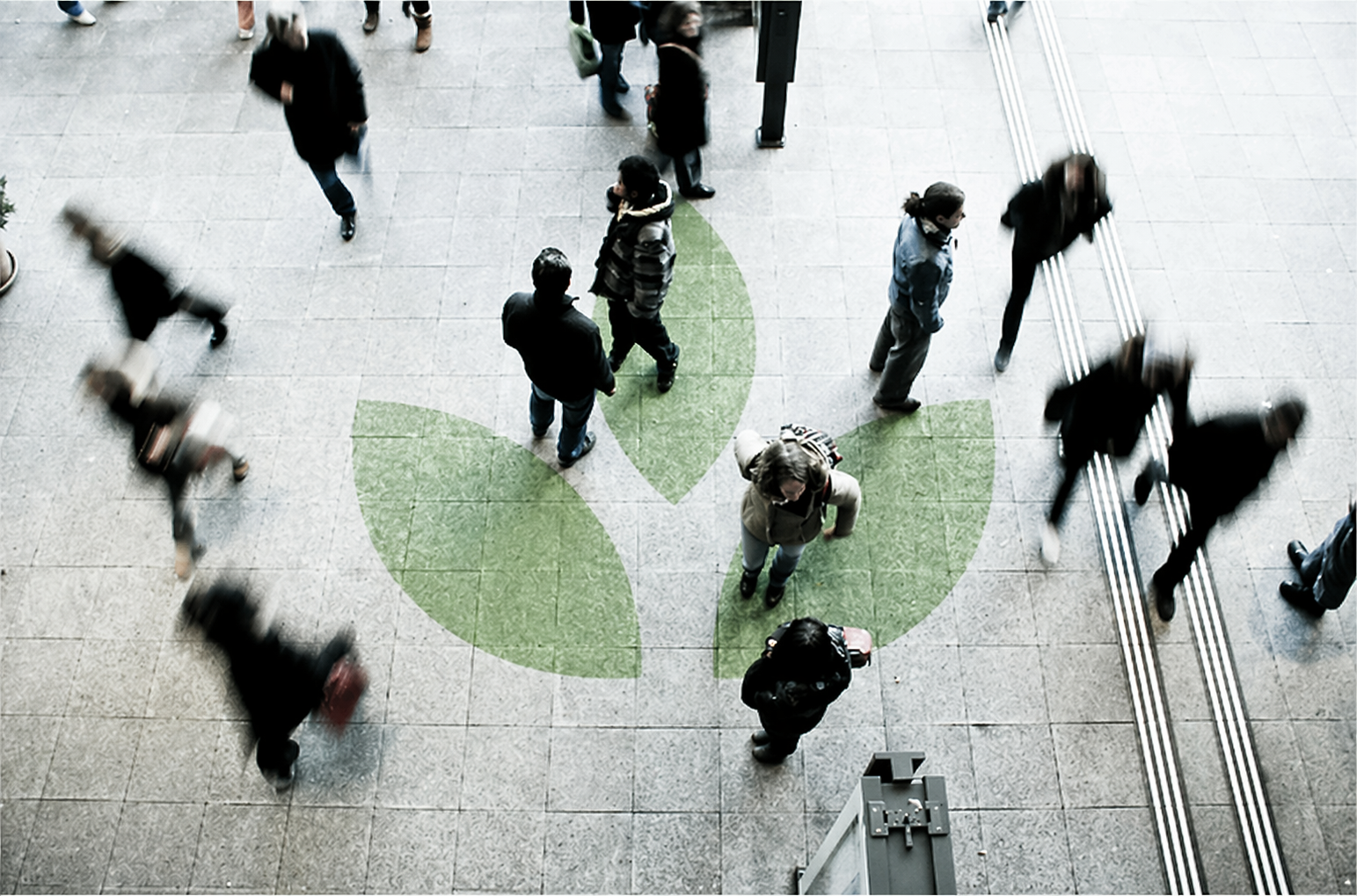 People walking on a tiled floor with green leaf-shaped designs, some blurred due to motion, at an indoor public space or transit station.