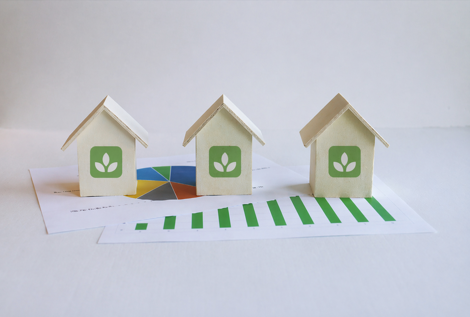 Three small white house models with green leaf logos on the front, placed on top of charts and reports, representing eco-friendly homes or sustainable living.