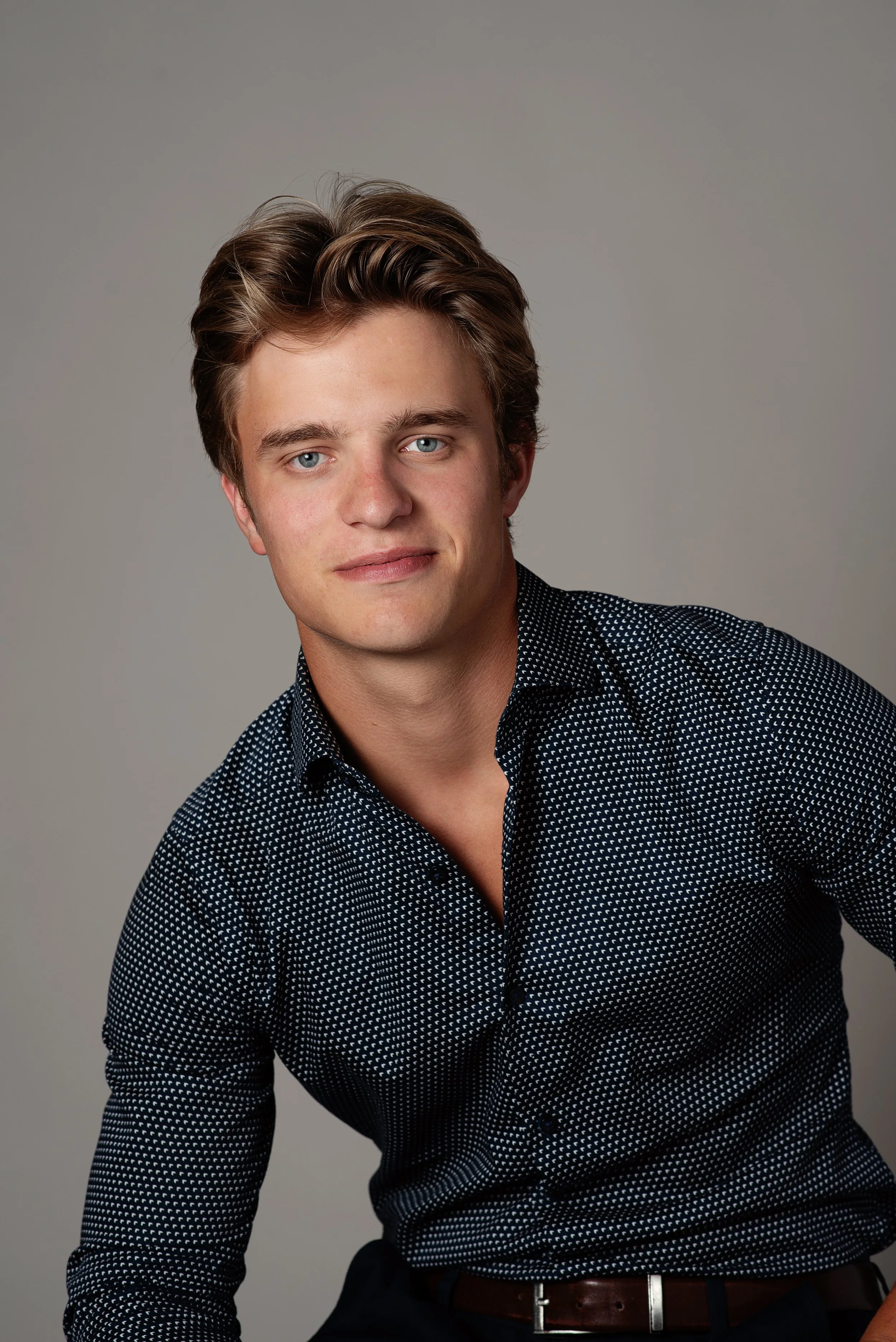 A young man with light skin, blue eyes, and styled brown hair, wearing a black and white polka dot shirt and a brown belt, looking at the camera with a slight smile against a neutral grey background.