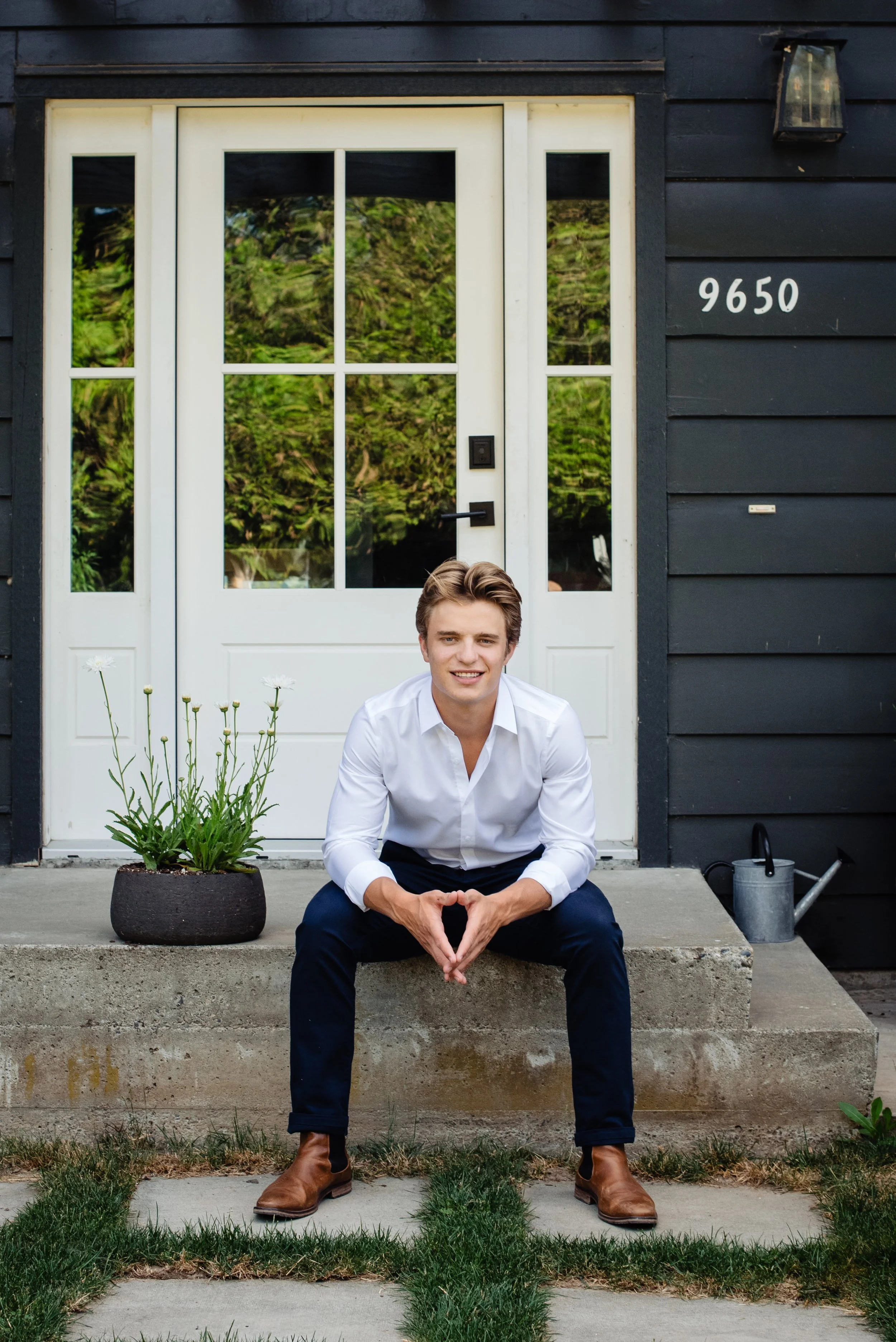 A young man with light brown hair, wearing a white shirt and dark pants, sitting on the front steps of a house with black siding and a white door, smiling at the camera.