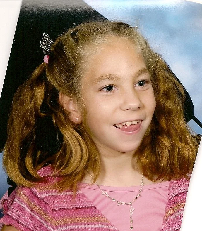 A young girl with wavy red hair, wearing a pink shirt and a necklace, smiling , sitting in a wheelchair. This is KiKi