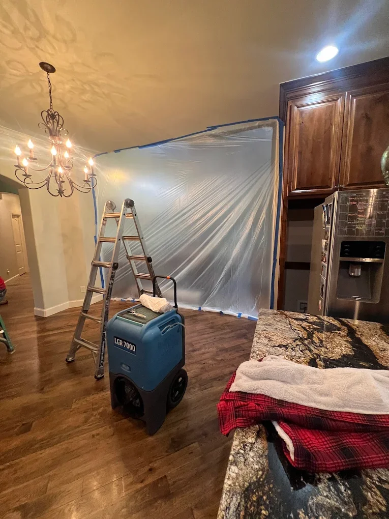 Kitchen and dining area with a plastic sheet hanging from the ceiling, a ladder, and a portable air scrubber, indicating ongoing renovations or painting.