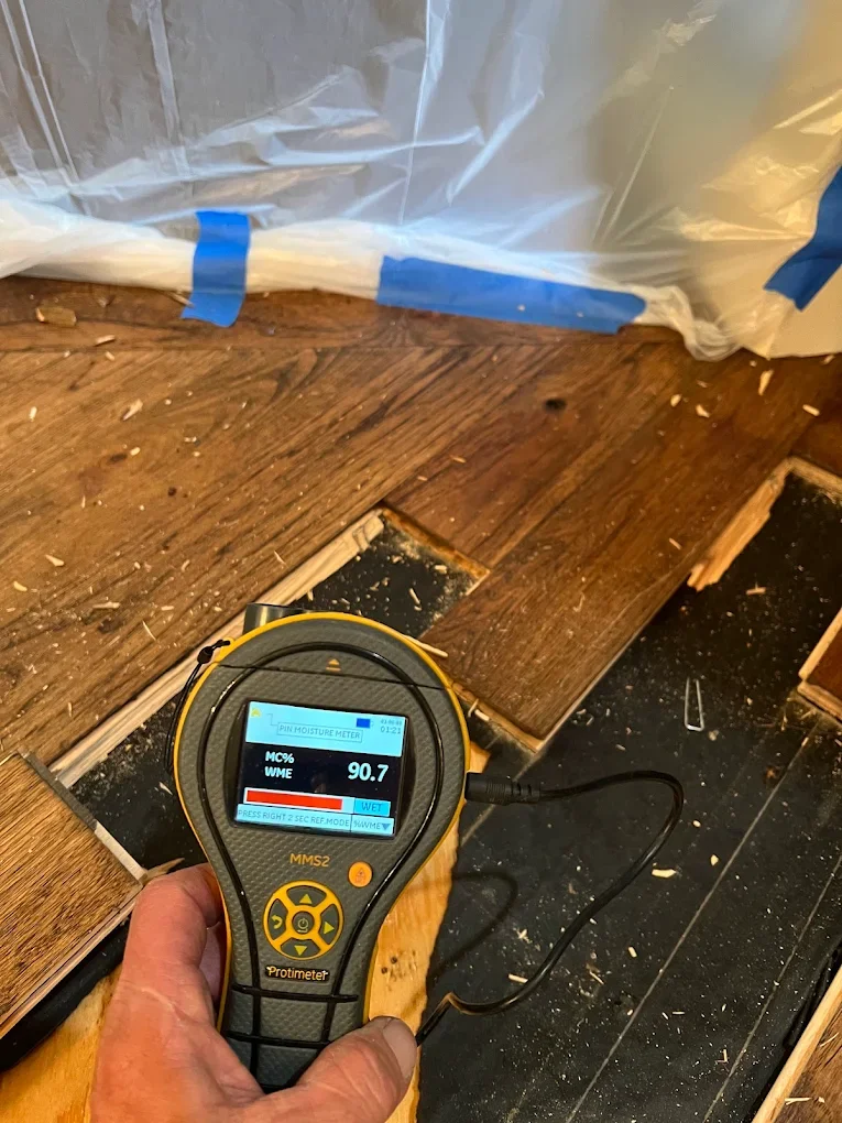 A moisture meter showing a reading of 90.7, being used on a wooden floor with a hole indicating water damage or rot.
