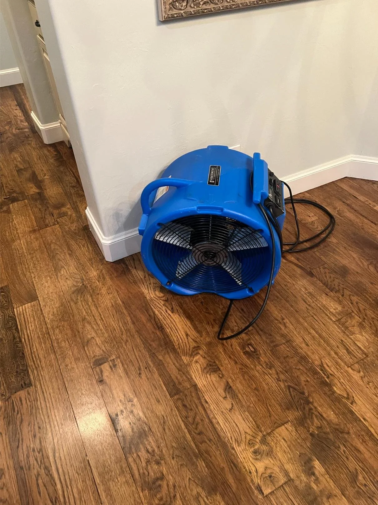 Blue industrial fan on a hardwood floor next to a white wall.