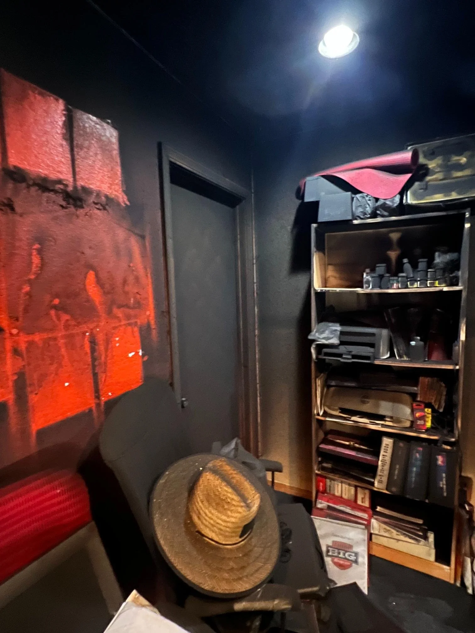 A cozy room with dark walls, a black bookshelf filled with books and items, a painting of red and black abstract art, a black chair, a woven hat on the chair, and a ceiling light.