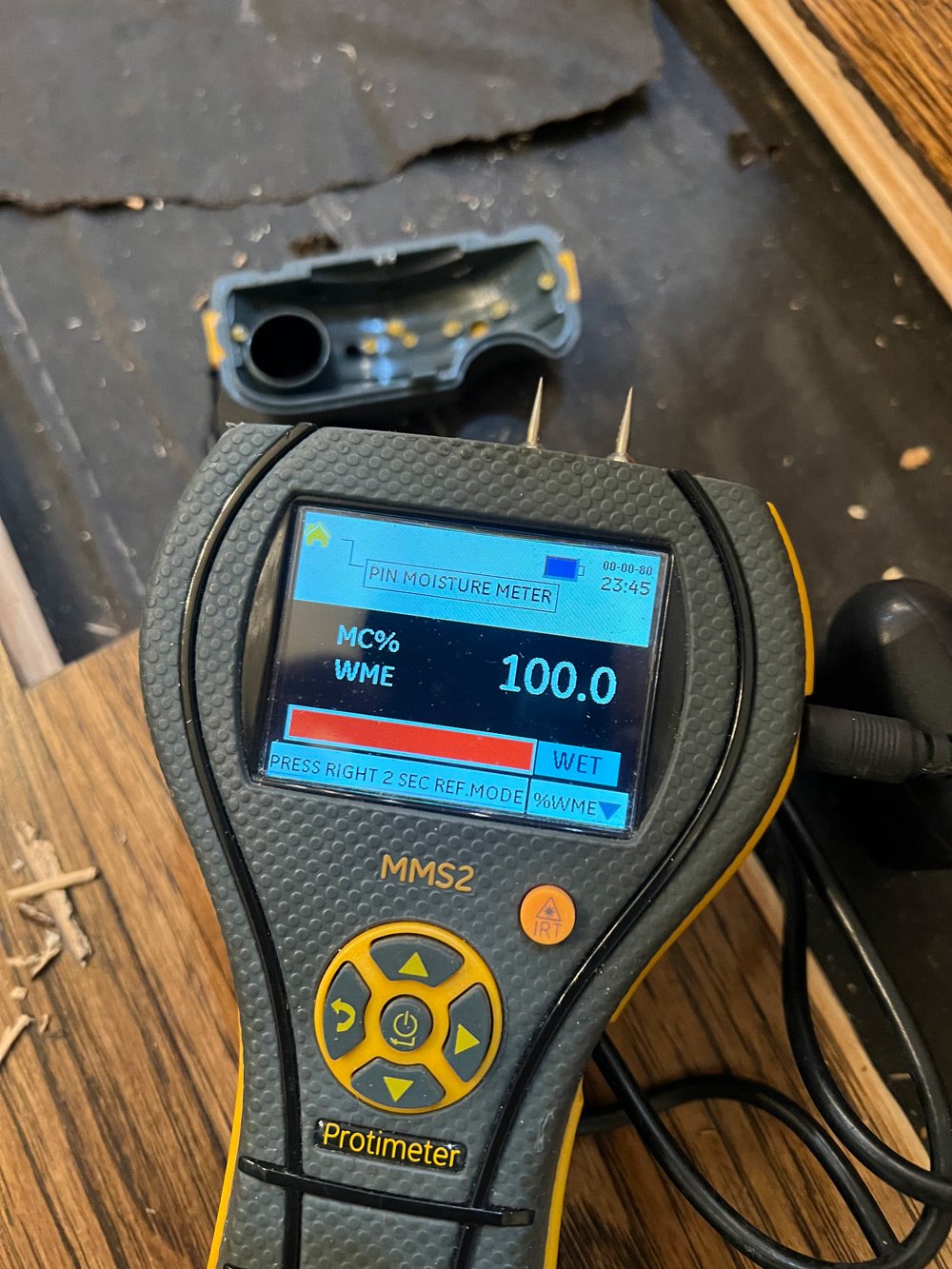 A digital pin moisture meter displaying 100.0% moisture content in the background on a wooden surface.