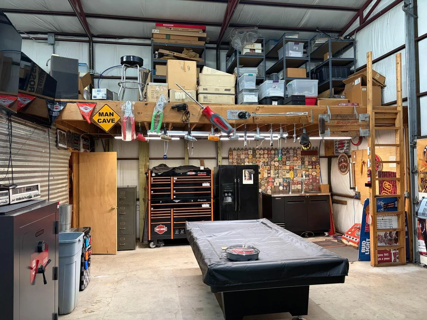 A garage or workshop area with a worktable covered in black plastic, tool storage cabinets, and pegboards with tools hanging. Shelves above hold boxes and supplies, with a ladder on the right. There are various signs, including a yellow "MAN CAVE" si