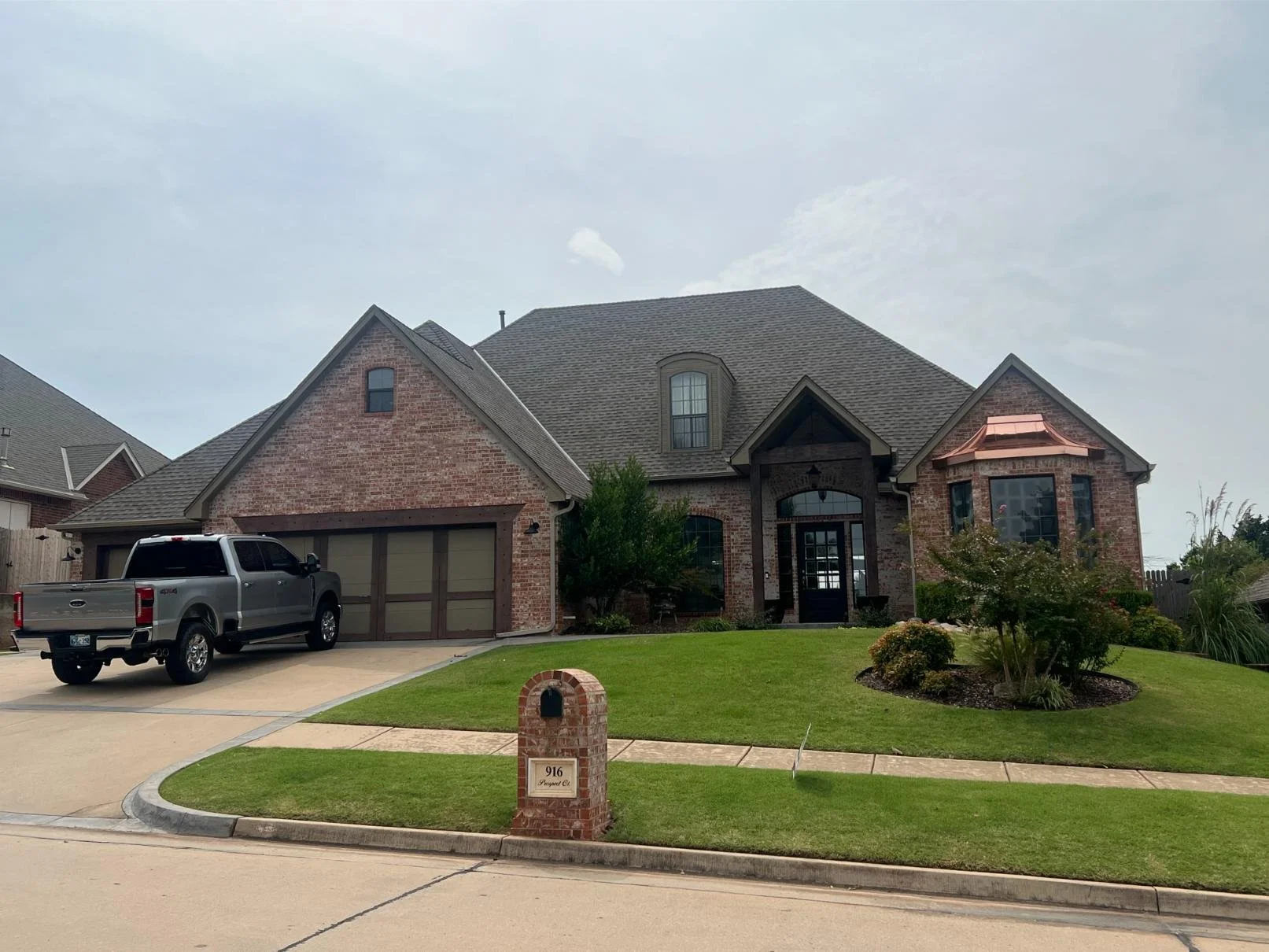 A two-story brick house with a garage, parked silver pickup truck in driveway, well-maintained lawn, bushes, and trees in front yard, mailbox with house number 916, and cloudy sky.