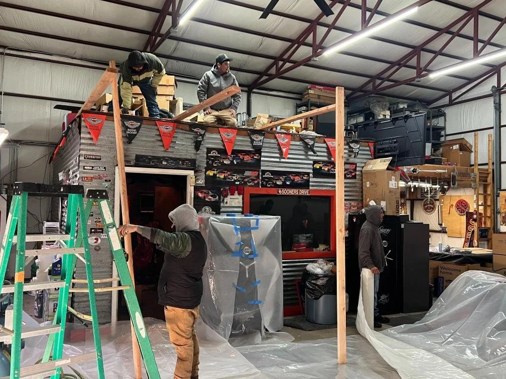 People constructing or renovating an indoor space, possibly a man in a hoodie and vest working with tools, another person on the roof installing wooden framework, and others observing or assisting amidst construction materials and equipment.