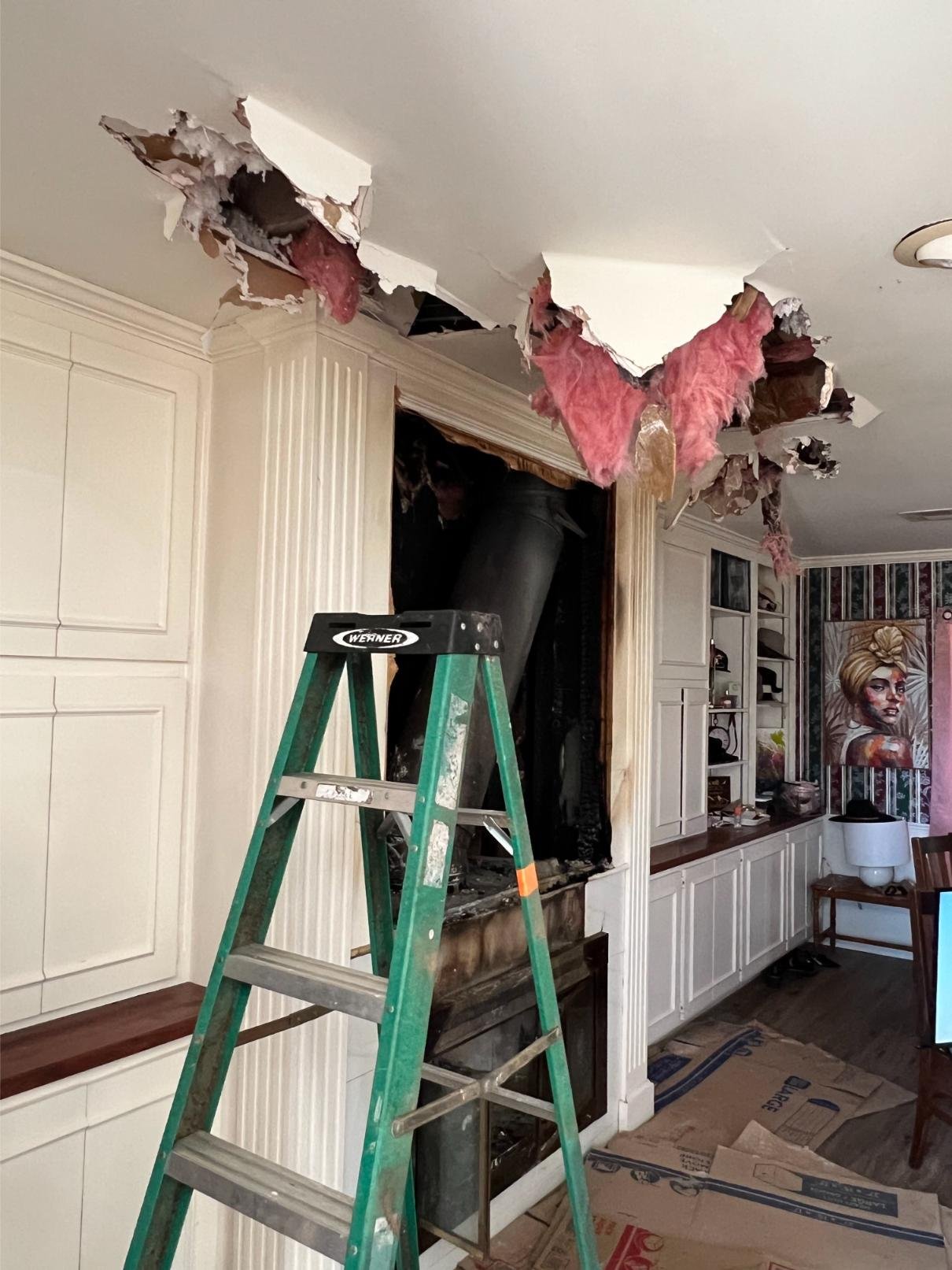 A living room with a partially collapsed ceiling above a fireplace, showing exposed insulation and debris. A green ladder stands in front, and there is artwork and shelves on the adjacent wall.