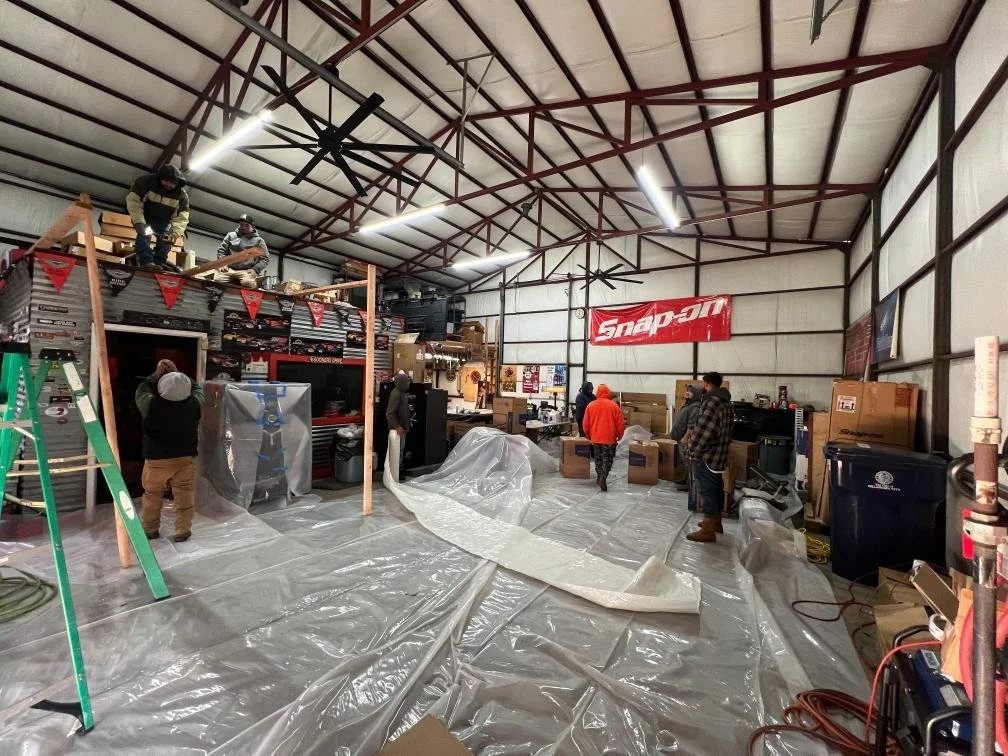 People working inside a large garage or workshop with a high ceiling, with tools, storage, and a red Snap-on banner. Some are assembling or arranging items, and a plastic sheet covers the floor.