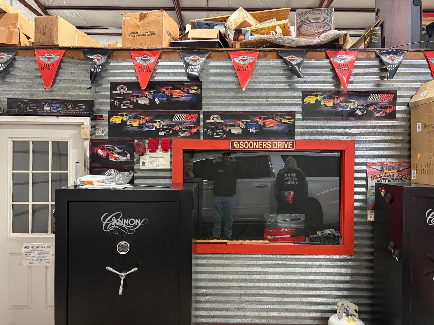 A garage wall decorated with racing car posters, red and black banners, and a mirror with a red frame labeled "Sooners Drive." There is a black safe and a metal cabinet with the label "Cannon". Several boxes and miscellaneous items are stored on top 