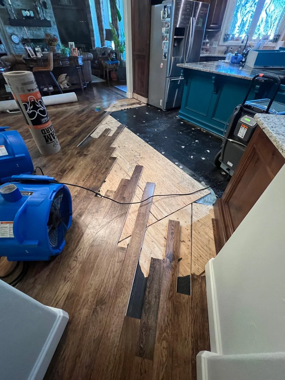 Partial removal of hardwood floor in a kitchen, with flooring pulled back and new flooring partially installed. Tools and construction equipment are visible, with a blue fan to dry water damage.