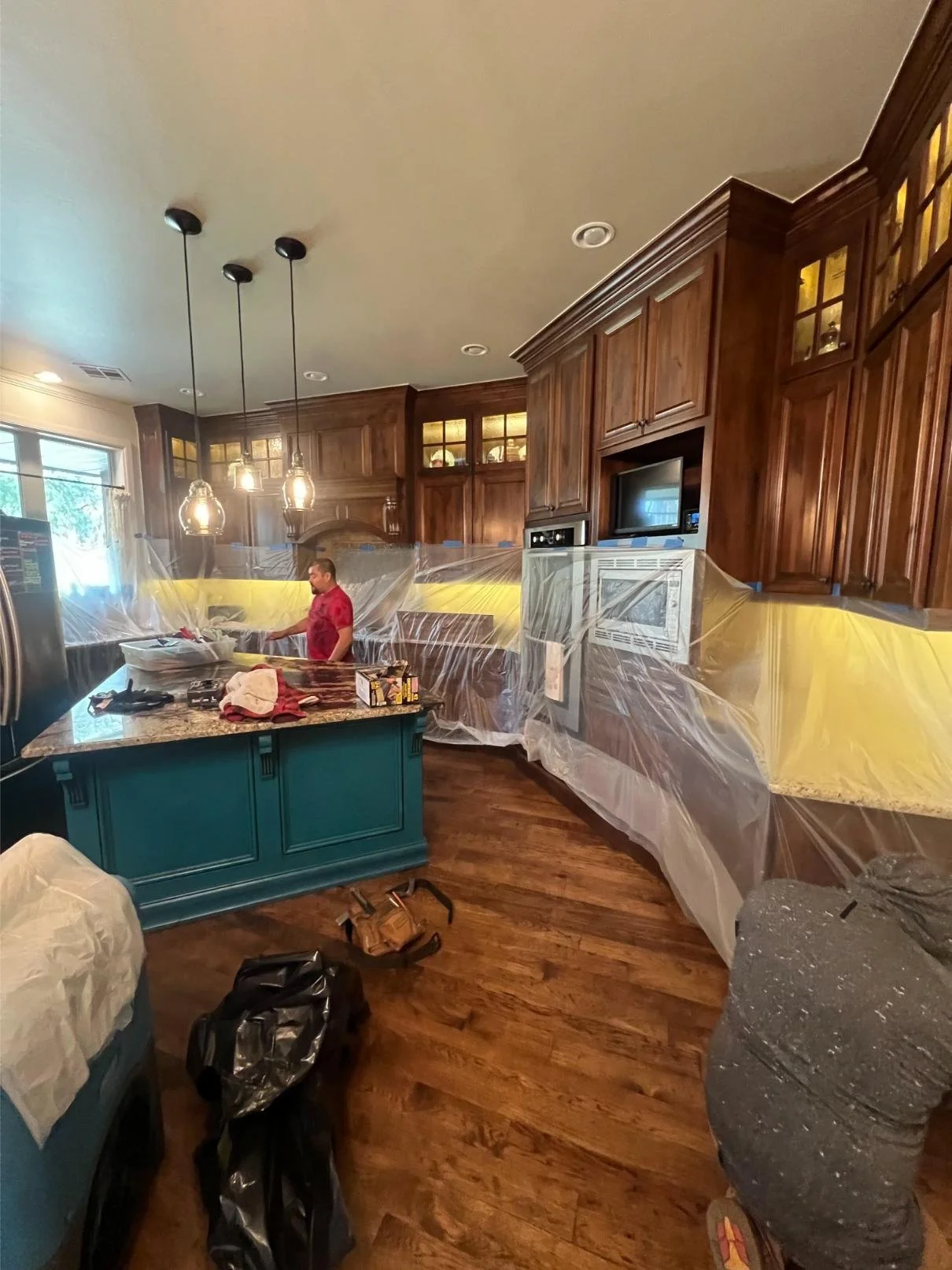 Kitchen undergoing renovation with plastic sheeting on cabinets and countertops, a man working at the counter, and various tools and clutter on the floor and island.