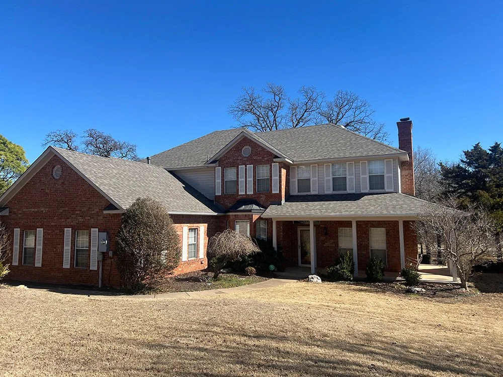 A large two-story brick house with a new roof, surrounded by few leafless trees and shrubs, under a clear blue sky.