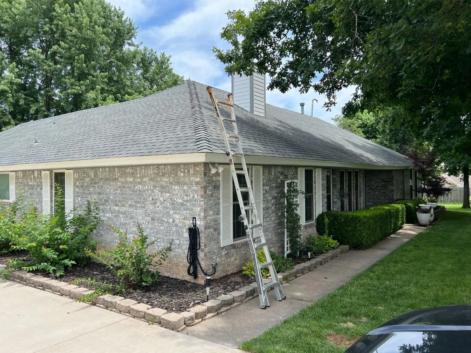 A house with a gray shingled roof and a chimney under a blue sky with some clouds. A ladder is leaned against the roof, possibly for maintenance or repair work. There is a garden with bushes along the side of the house, a sidewalk, and a grassy lawn 