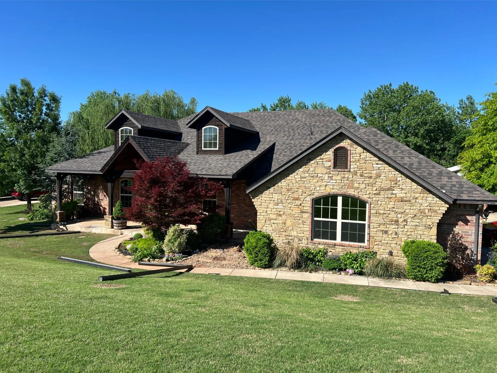 A large house with a mix of stone and brick exterior, multiple gabled roofs with dark shingles, large windows, and a manicured front yard with green grass, bushes, small trees, and a curved walkway.
