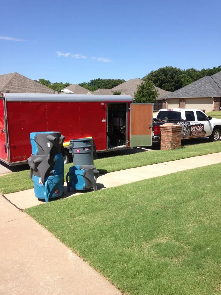 View of a residential driveway with a red trailer, two recycling bins, and a police vehicle with a Bulldog logo, in front of suburban houses under a blue sky.