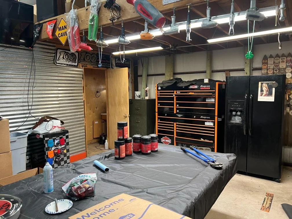 A cluttered garage with a table covered in a gray plastic tablecloth, cans, a blue wrapping paper roll, and a bottle on top. In the background, there are tool chests, a black refrigerator, a green filing cabinet, and various tools hanging from the ce