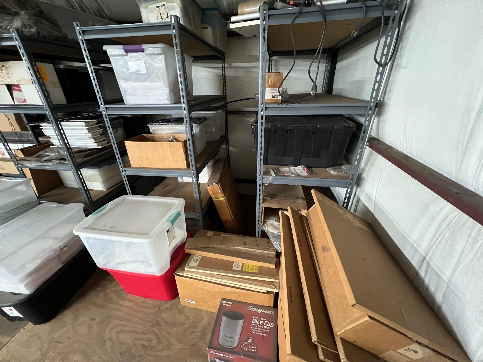 Garage storage shelves holding plastic bins, cardboard boxes, and miscellaneous items, with some empty space and a wooden piece on the floor.