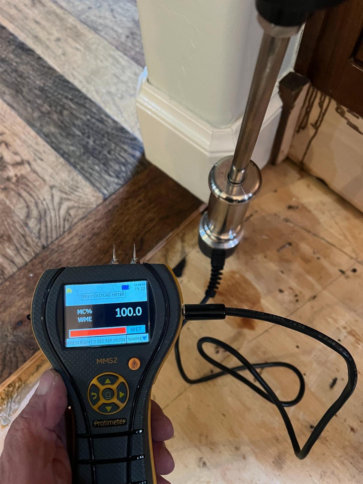 A moisture meter reading 100% wet on a vertical metal pipe or pole, with a background of wood and painted trim.