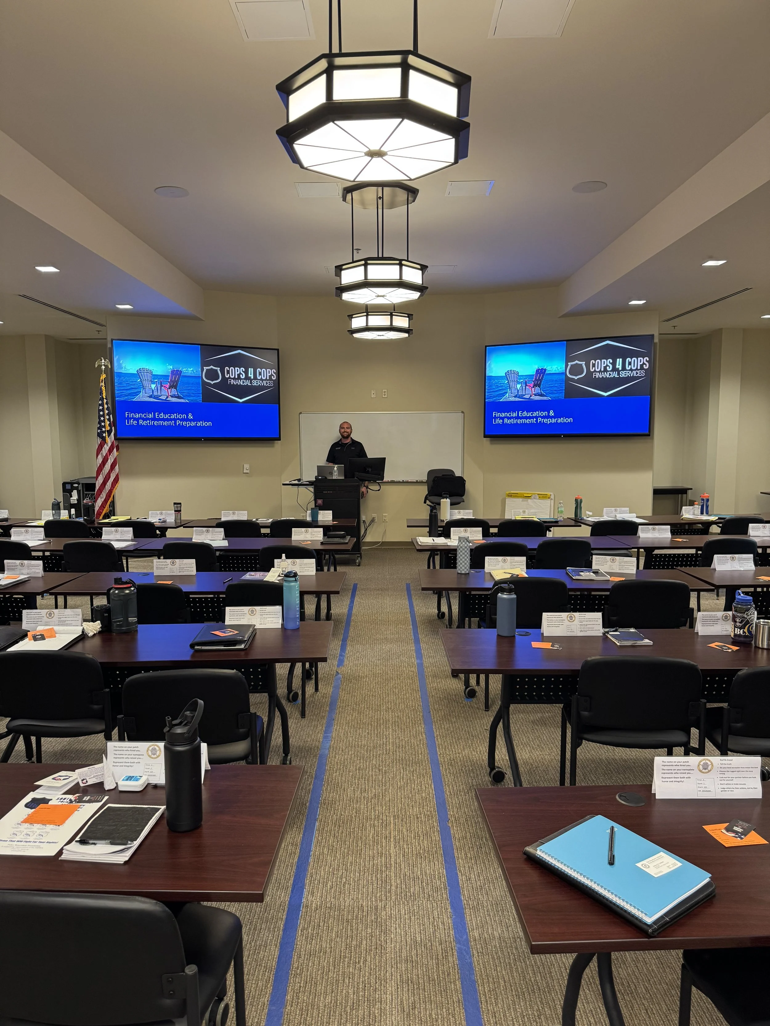 A classroom or conference room set up with tables and chairs, two large screens displaying a presentation titled 'COPS 4 COPS, Financial Services, Financial Education & Life Retirement Preparation,' and a man standing at a podium near a whiteboard. The room has ceiling lights, an American flag, water bottles, notebooks, and name tags on the tables.