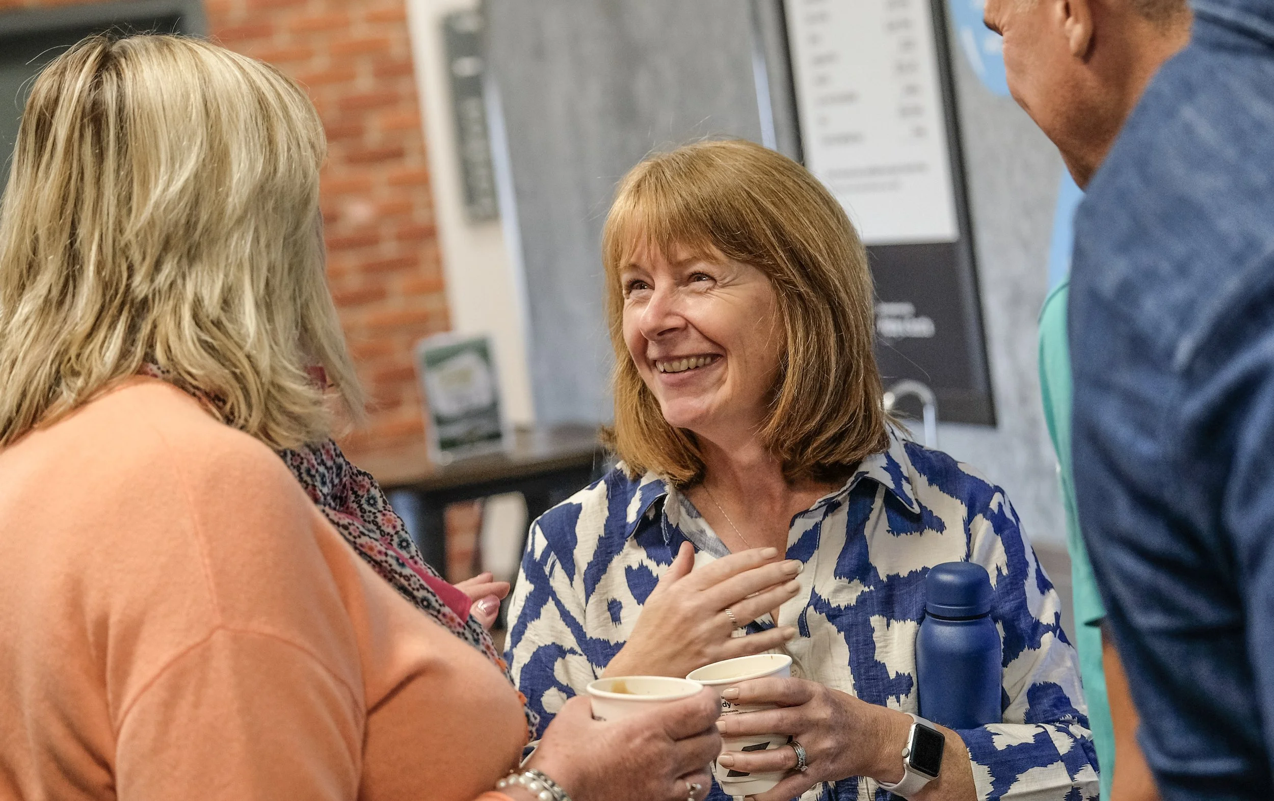Three women and a man engaged in conversation, with two women holding cups of coffee. The setting appears to be indoors, possibly at a social or professional gathering.
