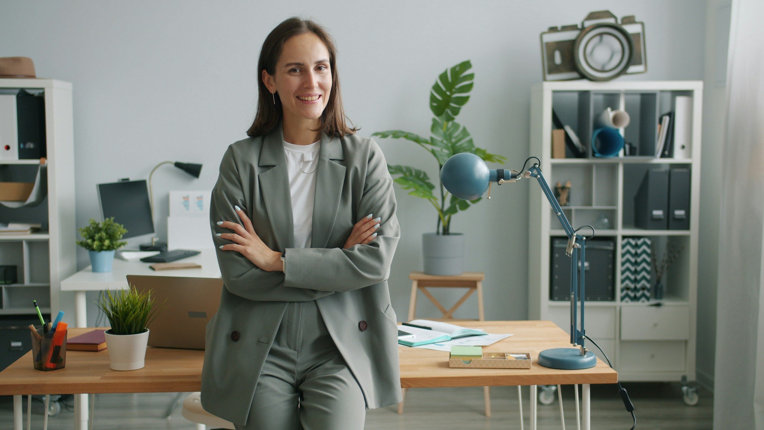 A woman with brown hair smiling, wearing a gray blazer and pants, standing with arms crossed in an office space with a desk, plants, shelves, and a camera on a shelf in the background.